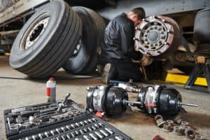 A mechanic from a mobile truck repair service is diligently working on the brake system of a large vehicle in a garage. Various tools and parts, including a toolbox and brake chambers, are scattered on the ground around him. The large tires of the vehicle are also visible in the background.
