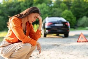 A woman in an orange shirt and beige pants kneels on the ground, holding her forehead in frustration. Behind her, a black car is parked on the side of a road with a red warning triangle placed on the ground. Mobile Truck Repair services are often needed to avoid such roadside dilemmas. Green trees and foliage fill the background.