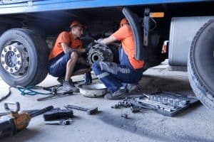 Two mechanics in orange shirts and dark shorts work on the rear wheel assembly of a large truck. Tools and parts, including a socket set and wrenches, are scattered around them on the ground. The scene takes place in a workshop specializing in mobile truck repair.