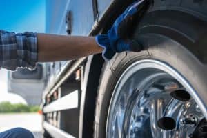 A person wearing gloves checks the tire of a large truck. The close-up image focuses on the shiny, reflective wheel and the person's hand touching the tire. The background shows part of the truck and a blurred outdoor scene, capturing a moment of skilled care in Mobile Truck Repair.