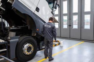 A mechanic in a grey uniform stands next to a raised truck cab in a large garage. There's a watering can on the floor nearby. The garage has large windows and doors in the background.